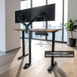 A modern sit-stand desk setup featuring dual monitors mounted on adjustable arms with the Cable Management Spine attached underneath, neatly organizing multiple blue cables from the desk surface to the floor. The flexible, snap-together segments protect and guide cables, connecting to a cable management tray under the desk, providing a clean, clutter-free workspace ideal for varied desk heights and active office environments.