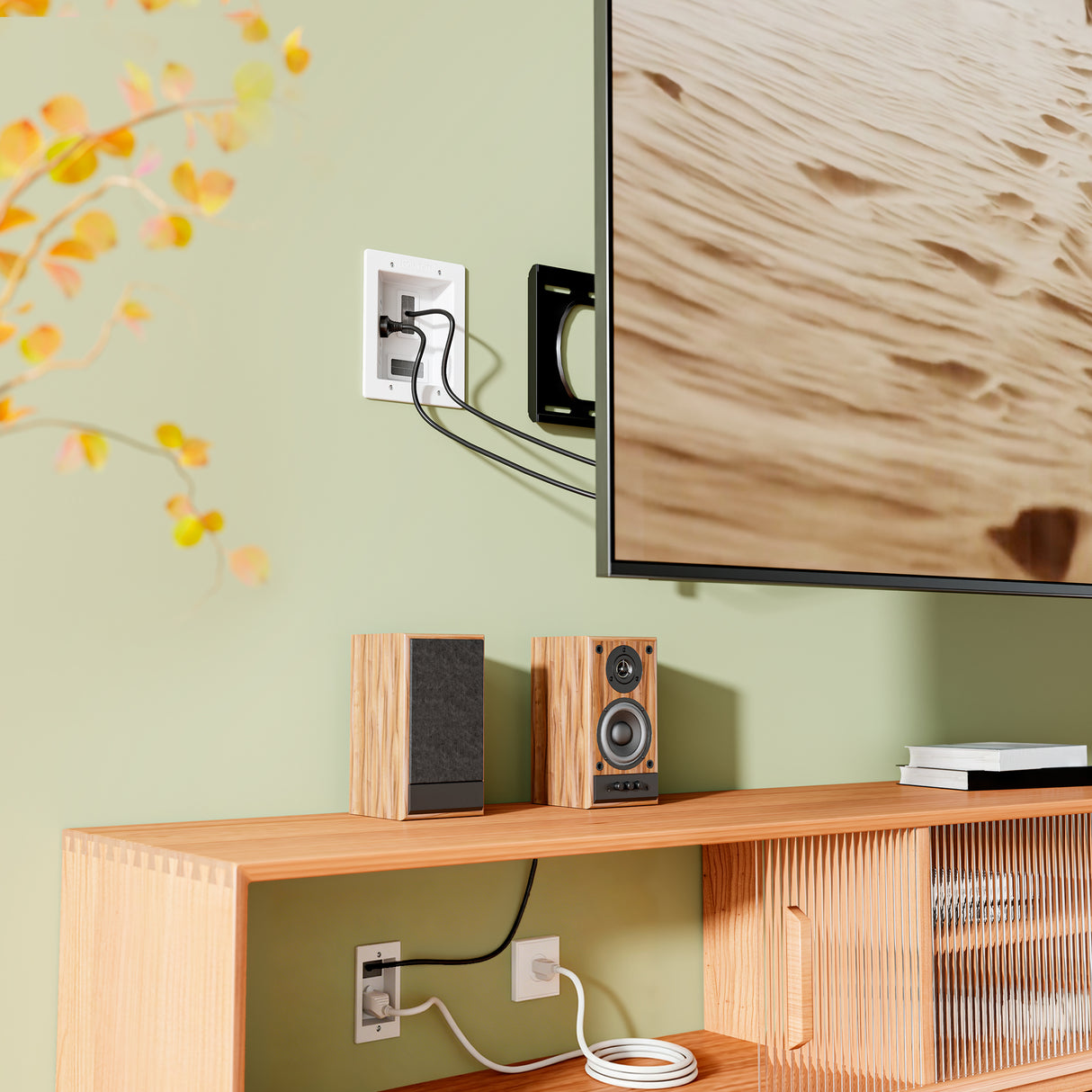 A wall-mounted TV with cords concealed by the Mount-It! In-Wall Cable Management Kit with USB and Power Input Module above a wooden media console holding speakers, books, and a coiled cable; a leafy branch appears on the left.