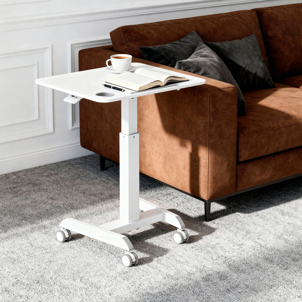 The Mount-It! Sit-Stand Mobile Laptop Cart, in white, serves as a versatile mobile workstation holding coffee and an open book before a brown sofa with dark pillows, all set atop a gray carpet in a bright room.