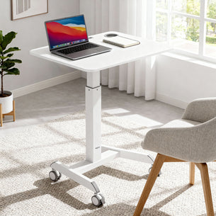 The Mount-It! Sit-Stand Mobile Laptop Cart, in white and height adjustable with wheels, holds a laptop and mouse. It stands in a bright room with large windows, white curtains, a light rug, and a gray cushioned chair with wooden legs.