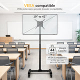 The Mount-It! Adjustable Mobile TV Stand with locking casters is set up in a classroom with desks and chairs. This mobile cart supports 19”–42” screens up to 44 lbs and is VESA compatible with various mounting patterns.